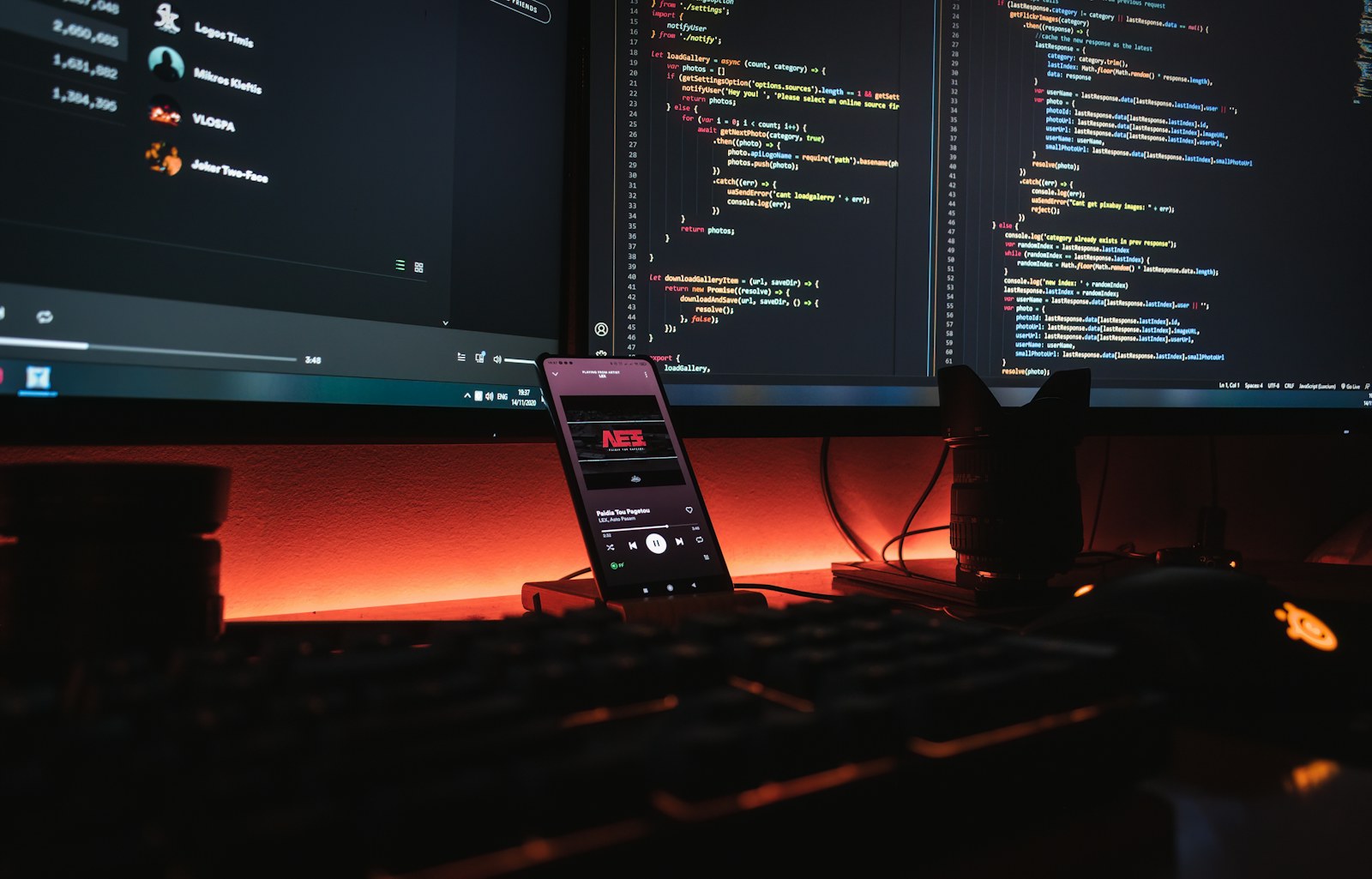 A smartphone displaying music on a desk with computer monitors showing code